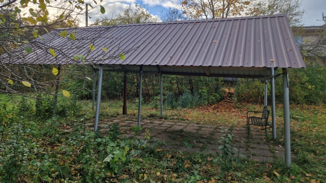 Metāla jumta nojume uz balstiem, zem tās bruģēts laukums un soliņš