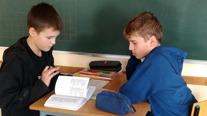 Divi puiši pie skolas sola ar mācību piederumiem un grāmatu