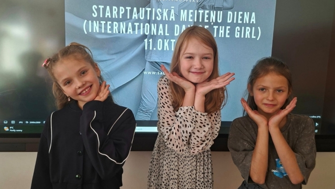 Trīs meitenes pozē Starptautiskajai meiteņu dienai veltīta plakāta priekšā