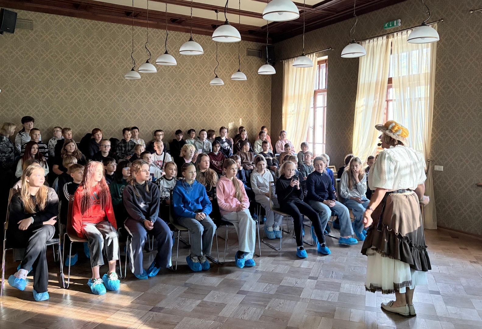 Skolēni klausās gides stāstījumu vēsturiskā telpā