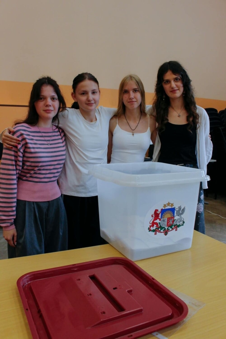 Četras jaunietes stāv pie vēlēšanu urnas ar Latvijas ģerboni