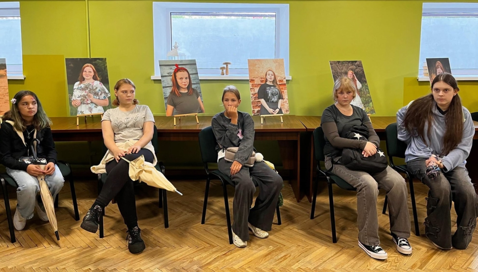 Meitenes sēž bibliotēkā, aiz viņām redzami izstādes portreti