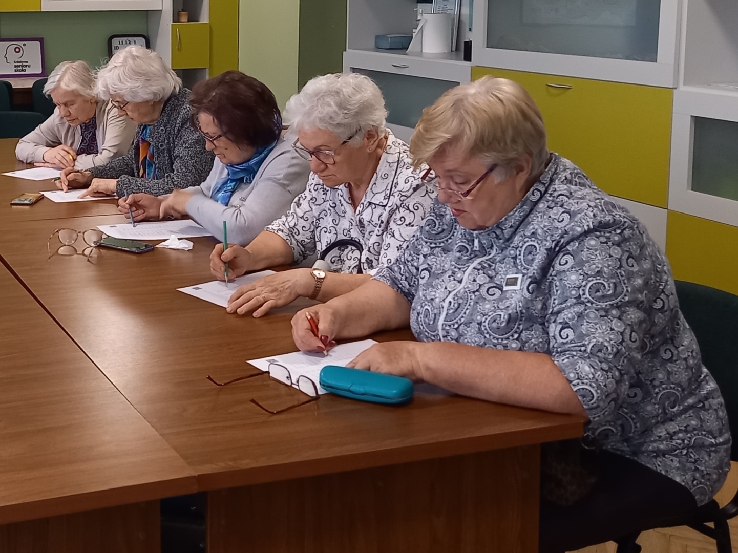Seniori sēž pie galda un pilda rakstisku uzdevumu