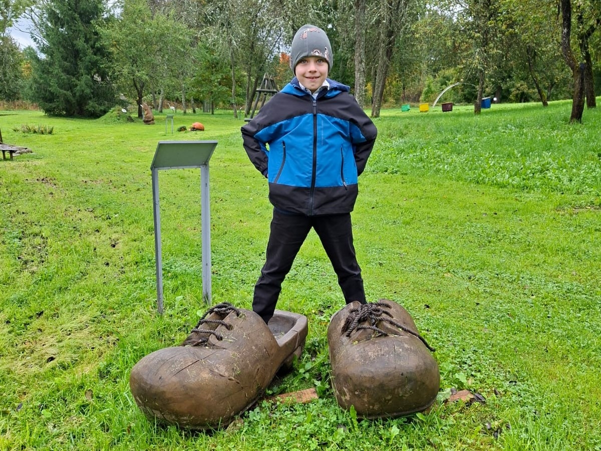 Skolēns pozē dārzā pie lieliem koka apaviem