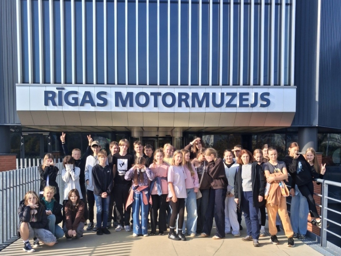 Skolēnu grupa kopīgi fotografējas pie Rīgas Motormuzeja ieejas