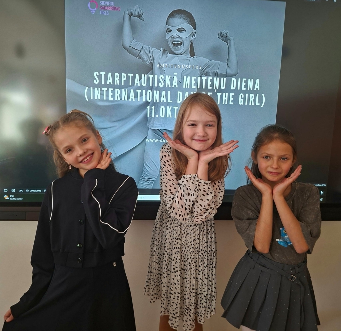Trīs meitenes pozē Starptautiskajai meiteņu dienai veltīta plakāta priekšā