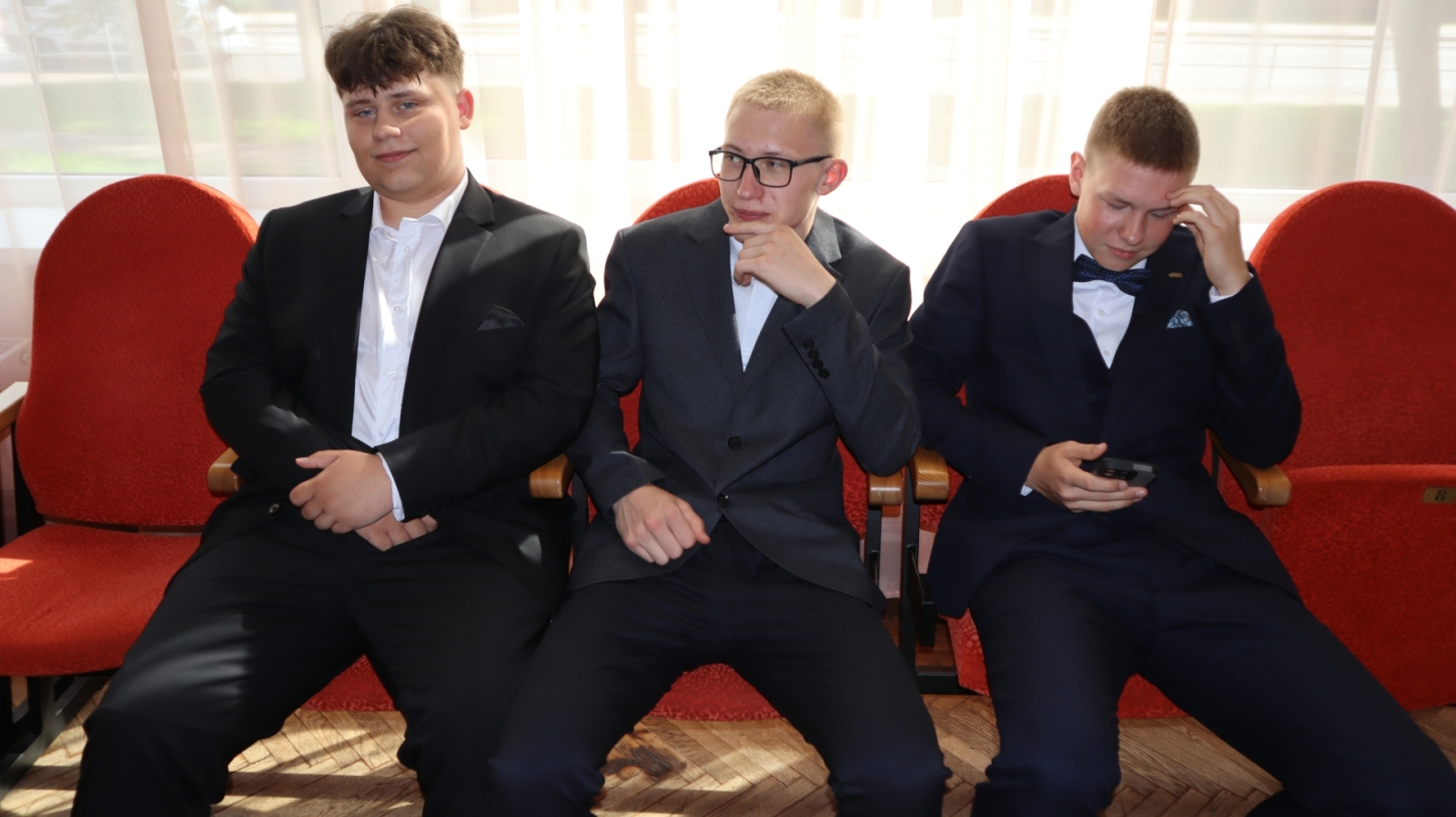 Trīs absolventi sēž krēslos pirms ceremonijas