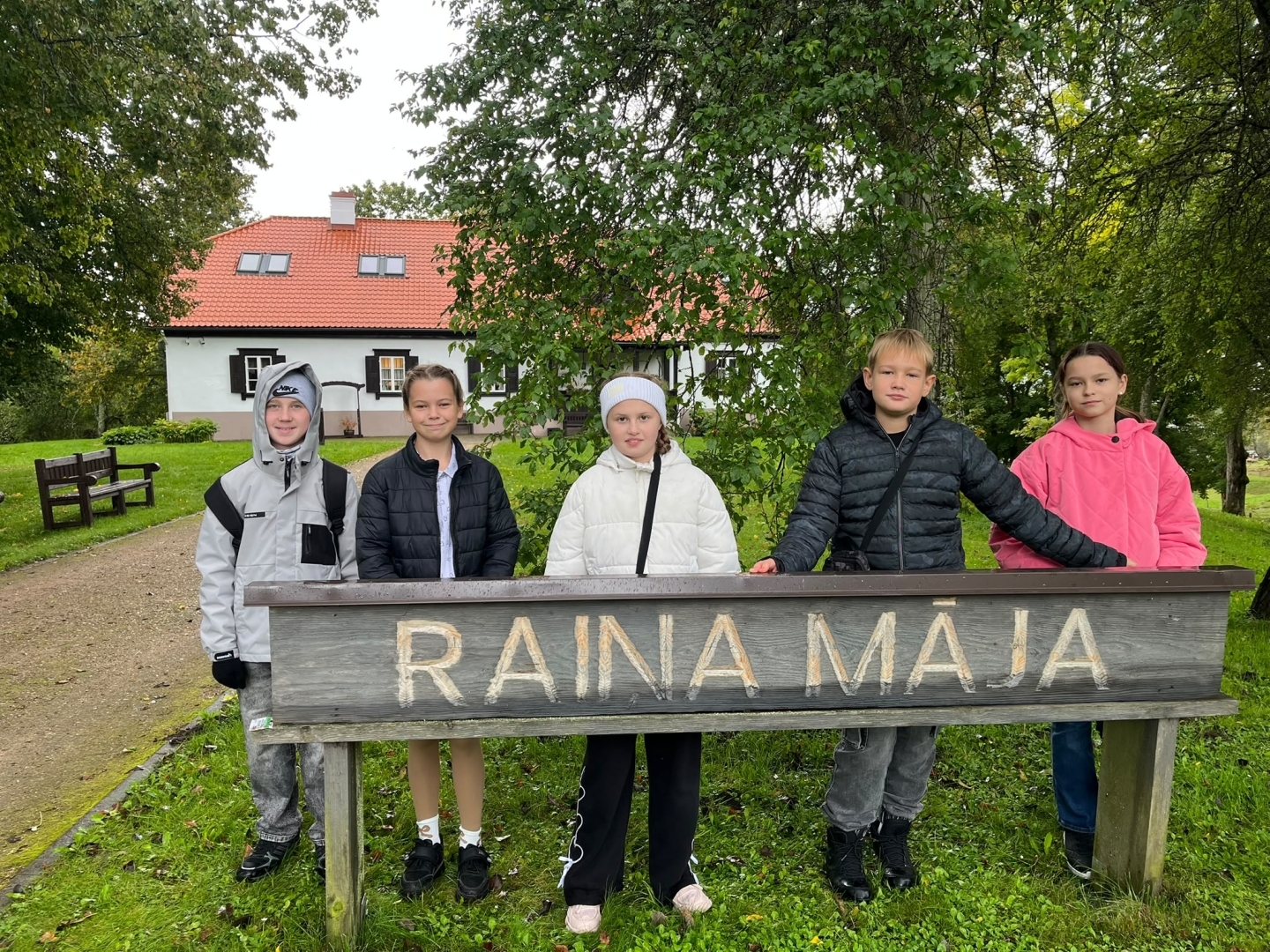 Pieci bērni stāv pie koka norādes „Raiņa Māja”