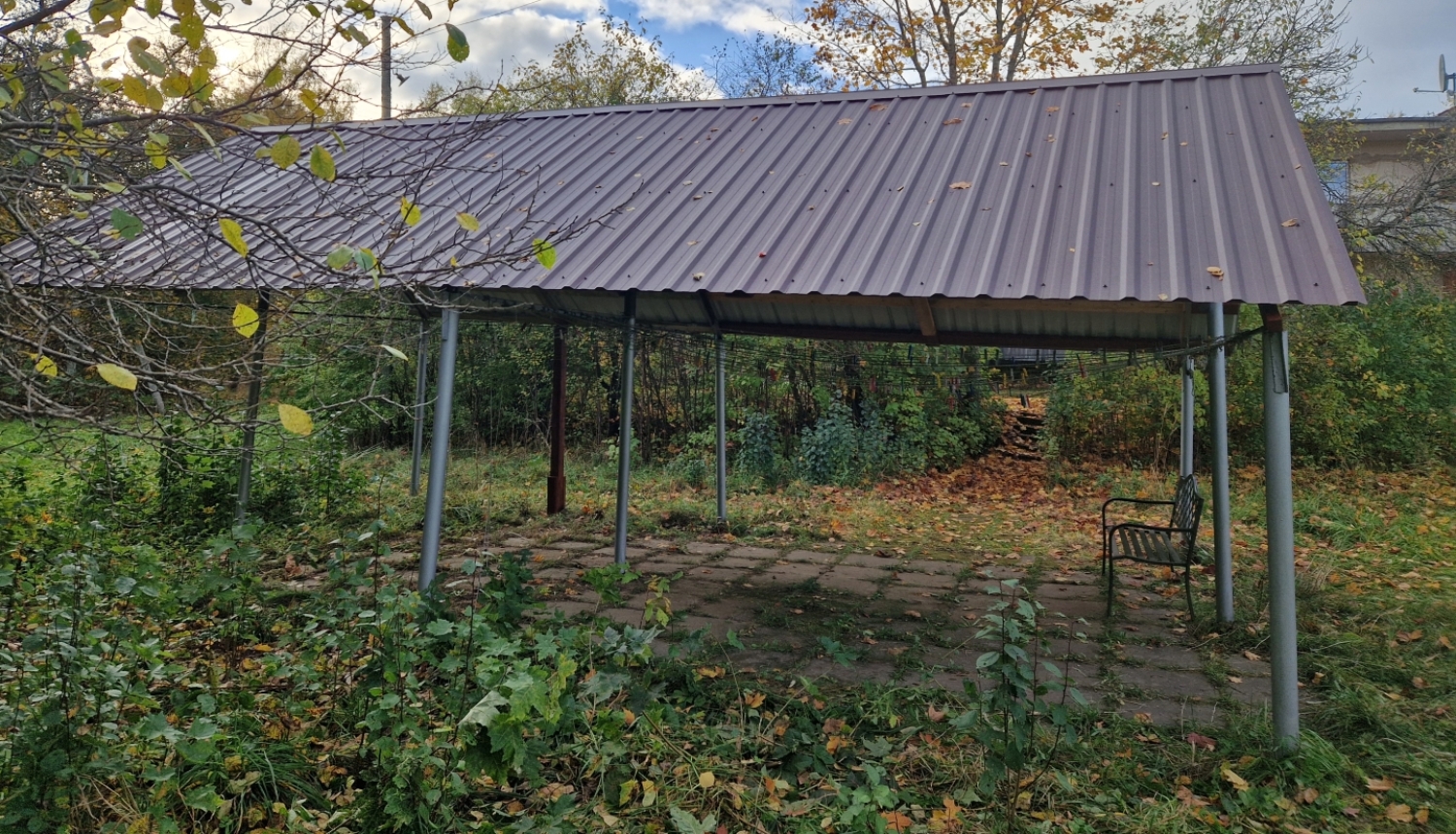 Metāla jumta nojume uz balstiem, zem tās bruģēts laukums un soliņš