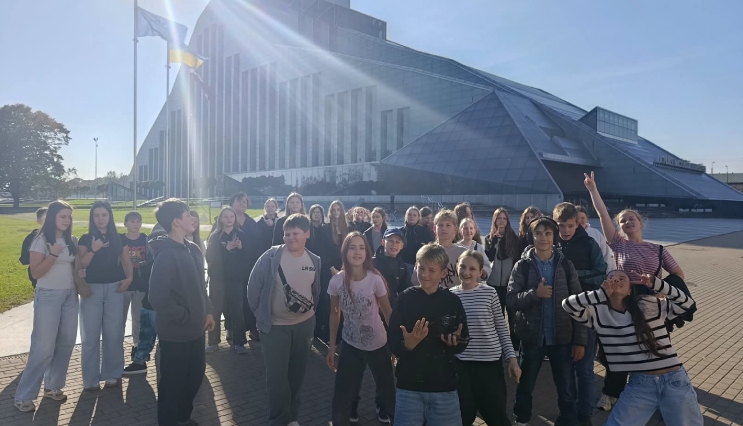 Skolēnu grupa pozē pie Latvijas Nacionālās bibliotēkas ēkas