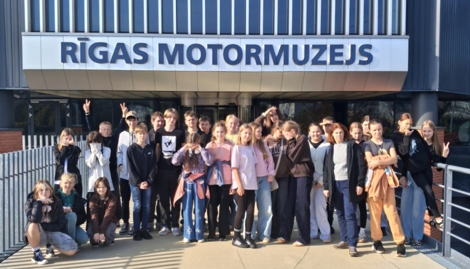 Skolēnu grupa kopīgi fotografējas pie Rīgas Motormuzeja ieejas