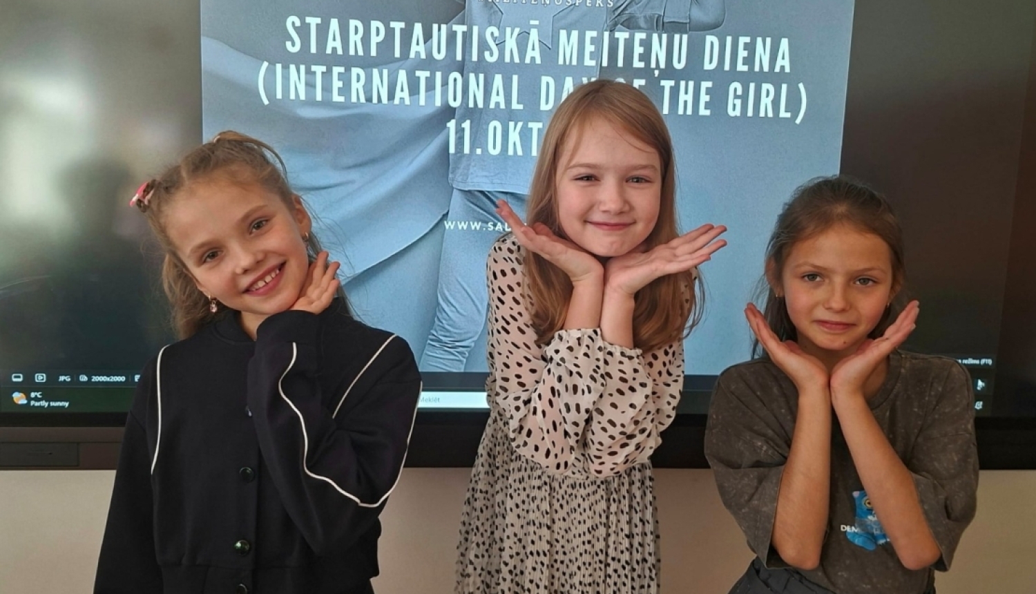 Trīs meitenes pozē Starptautiskajai meiteņu dienai veltīta plakāta priekšā
