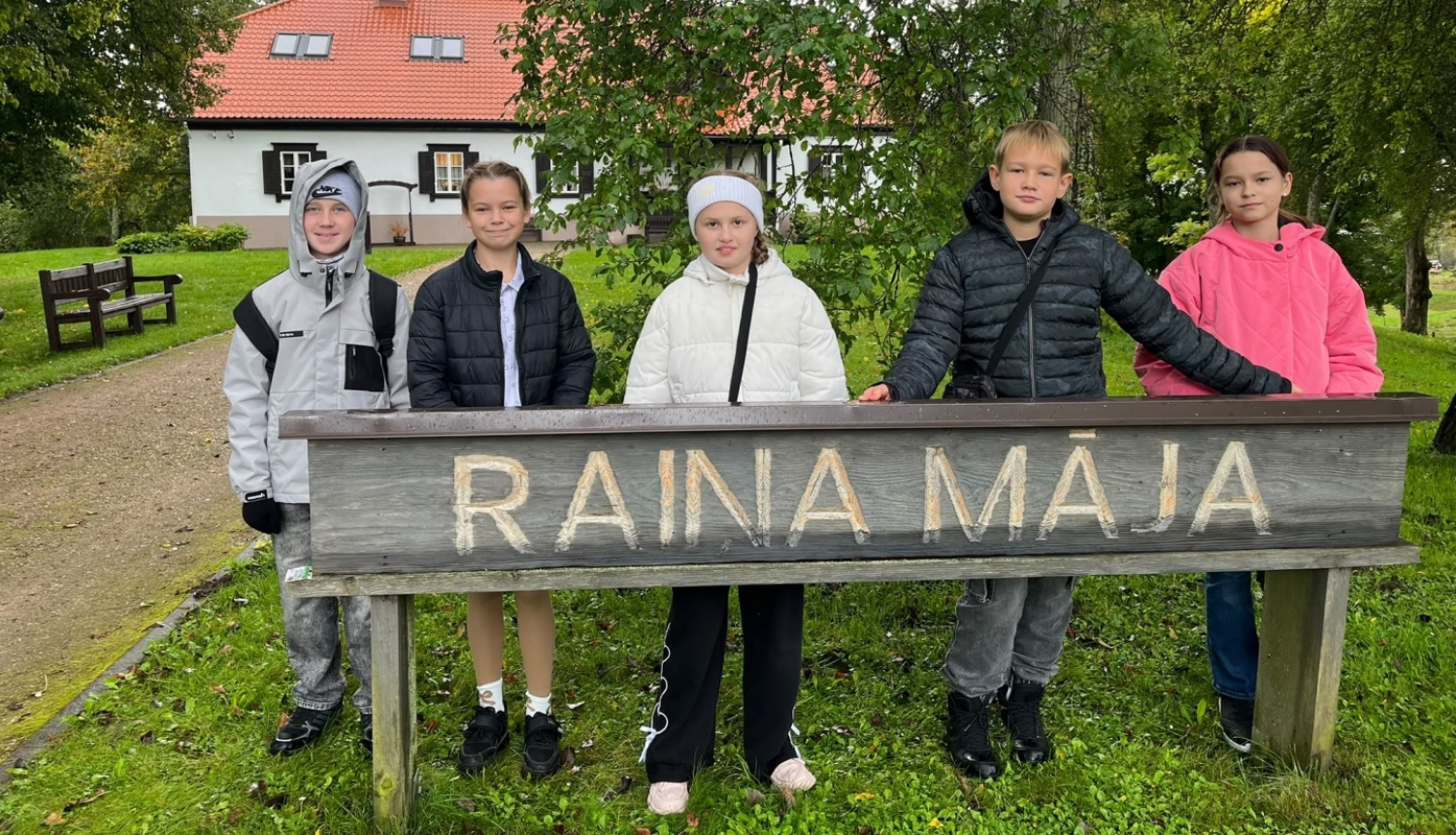 Pieci bērni stāv pie koka norādes „Raiņa Māja”