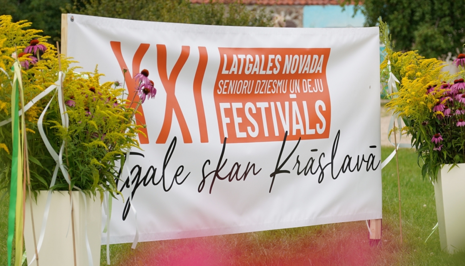 Senioru dziesmu un deju festivāla baneris dabā pie ziedu kompozīcijas