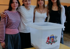 Četras jaunietes stāv pie vēlēšanu urnas ar Latvijas ģerboni