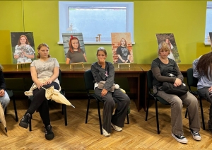 Meitenes sēž bibliotēkā, aiz viņām redzami izstādes portreti