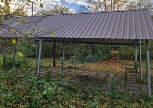 Metāla jumta nojume uz balstiem, zem tās bruģēts laukums un soliņš