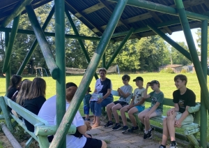 Bērni un skolotāja sēž zem koka nojumes un piedalās kopīgā nodarbībā