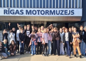 Skolēnu grupa kopīgi fotografējas pie Rīgas Motormuzeja ieejas