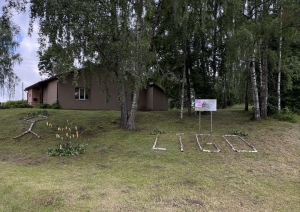 Zālienā no bērza stumbriem veidots uzraksts “LĪGO” pie ēkas un bērzu birzs
