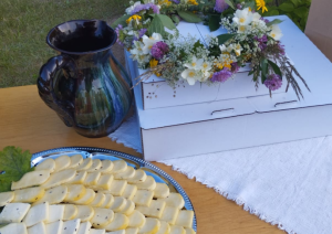 Uz galda izkārtotas siera šķēles, keramikas krūze un vasaras ziedu vainags