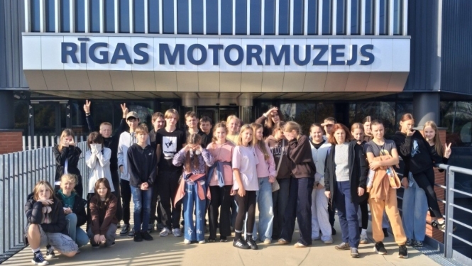 Skolēnu grupa kopīgi fotografējas pie Rīgas Motormuzeja ieejas
