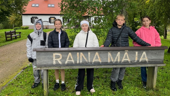 Pieci bērni stāv pie koka norādes „Raiņa Māja”